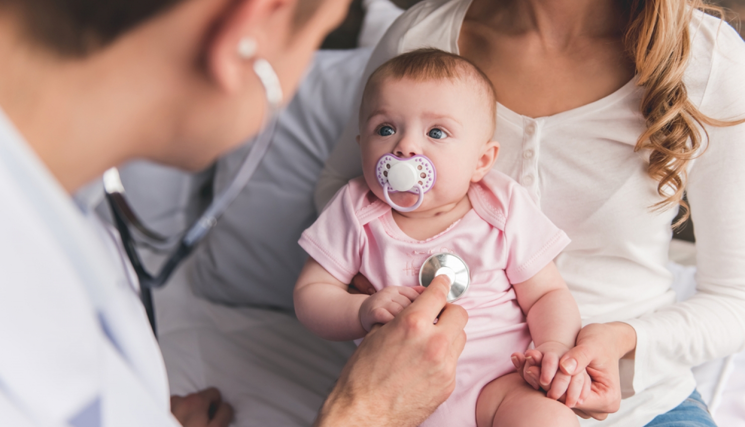 Ģimenes ārsts pārbauda mazuli ar stetoskopu. Mazulis ir rokās mammai. 